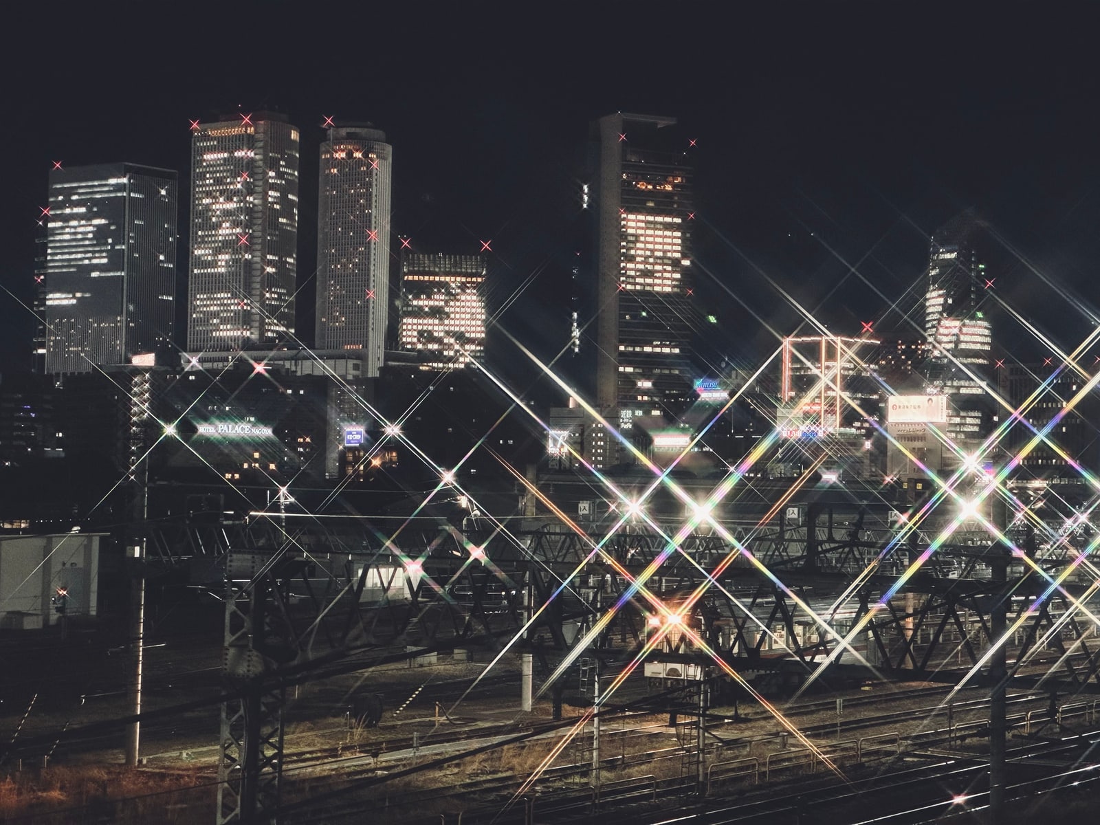 Lit towers behind a railway yard at night, a HOTEL PALACE NAGOYA sign at left, the lights spread into star shapes