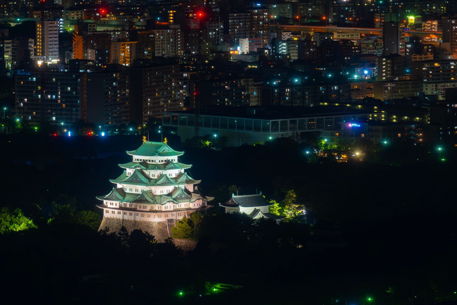 A lit castle keep with green copper roofs and gold ridge ornaments among dark trees at night, apartment blocks and city lights behind