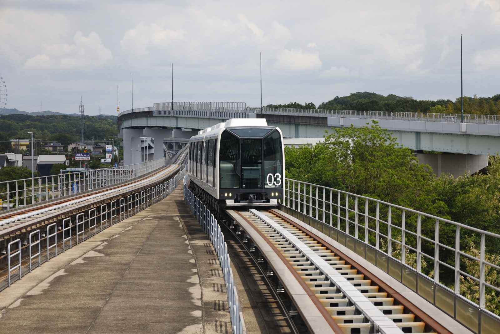 A short white and black train on an elevated guideway curving through green hills under an overcast sky