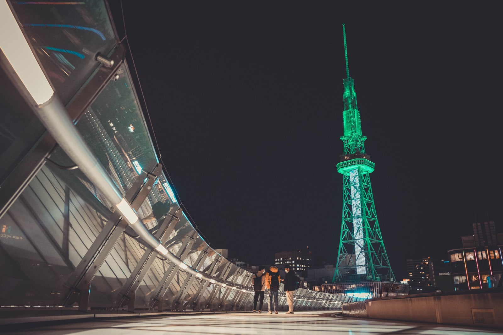 A lattice tower lit green at night seen from a curved glass-sided walkway, three people standing in the foreground