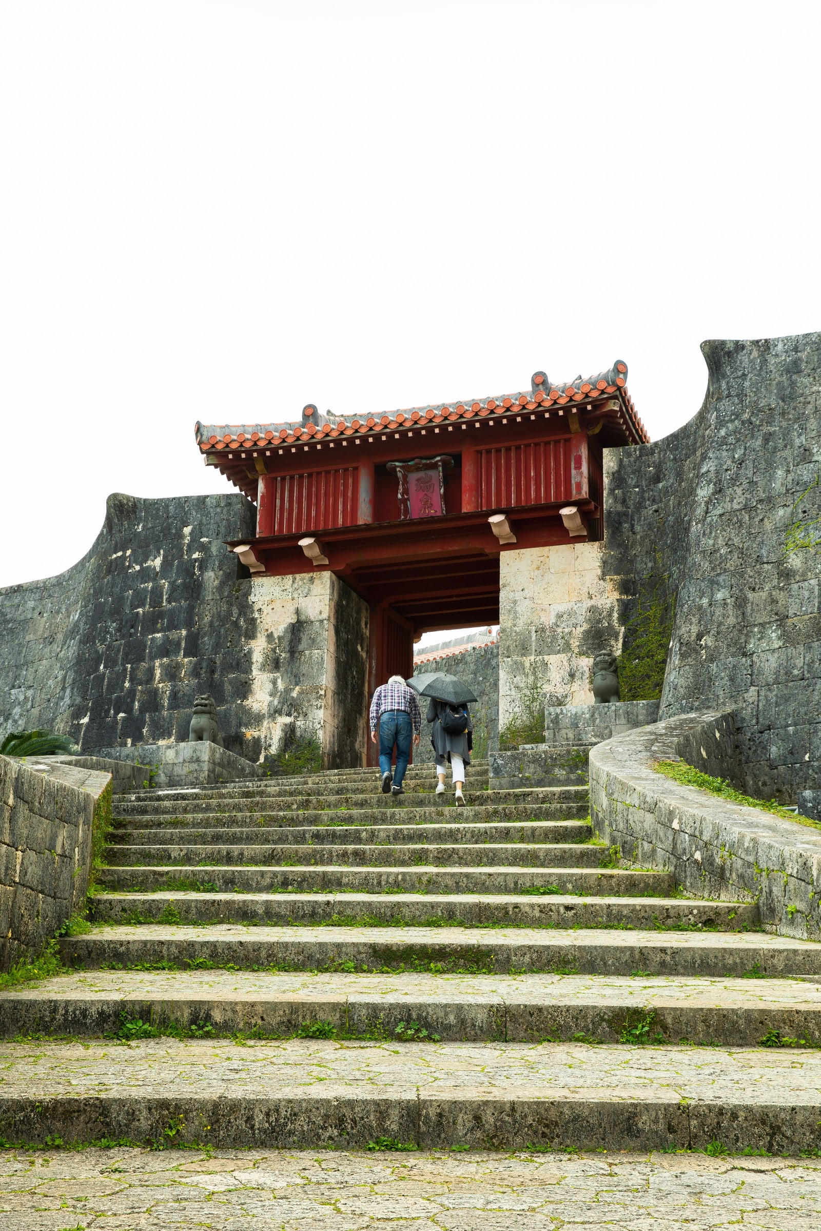 A red gate at the top of wide stone steps between stone walls at Shuri Castle Park, two visitors walking up, one under a black umbrella