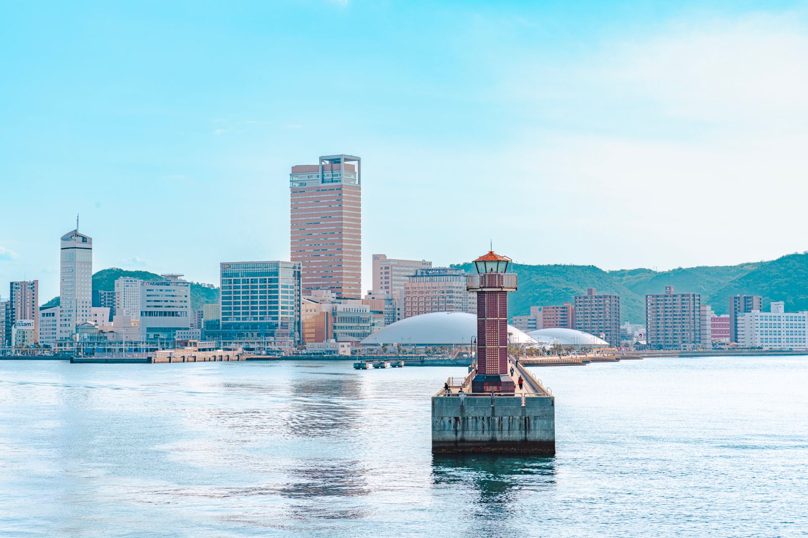 A red lighthouse on a breakwater, with the Takamatsu waterfront and green hills behind