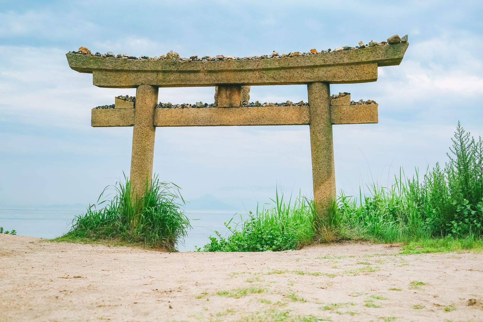 A stone torii gate on sand with pebbles stacked along its beams and the sea behind