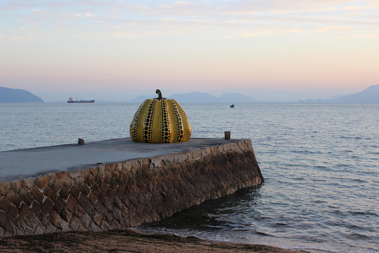 A yellow dotted pumpkin sculpture on a stone pier at dusk, a cargo ship far out on calm sea