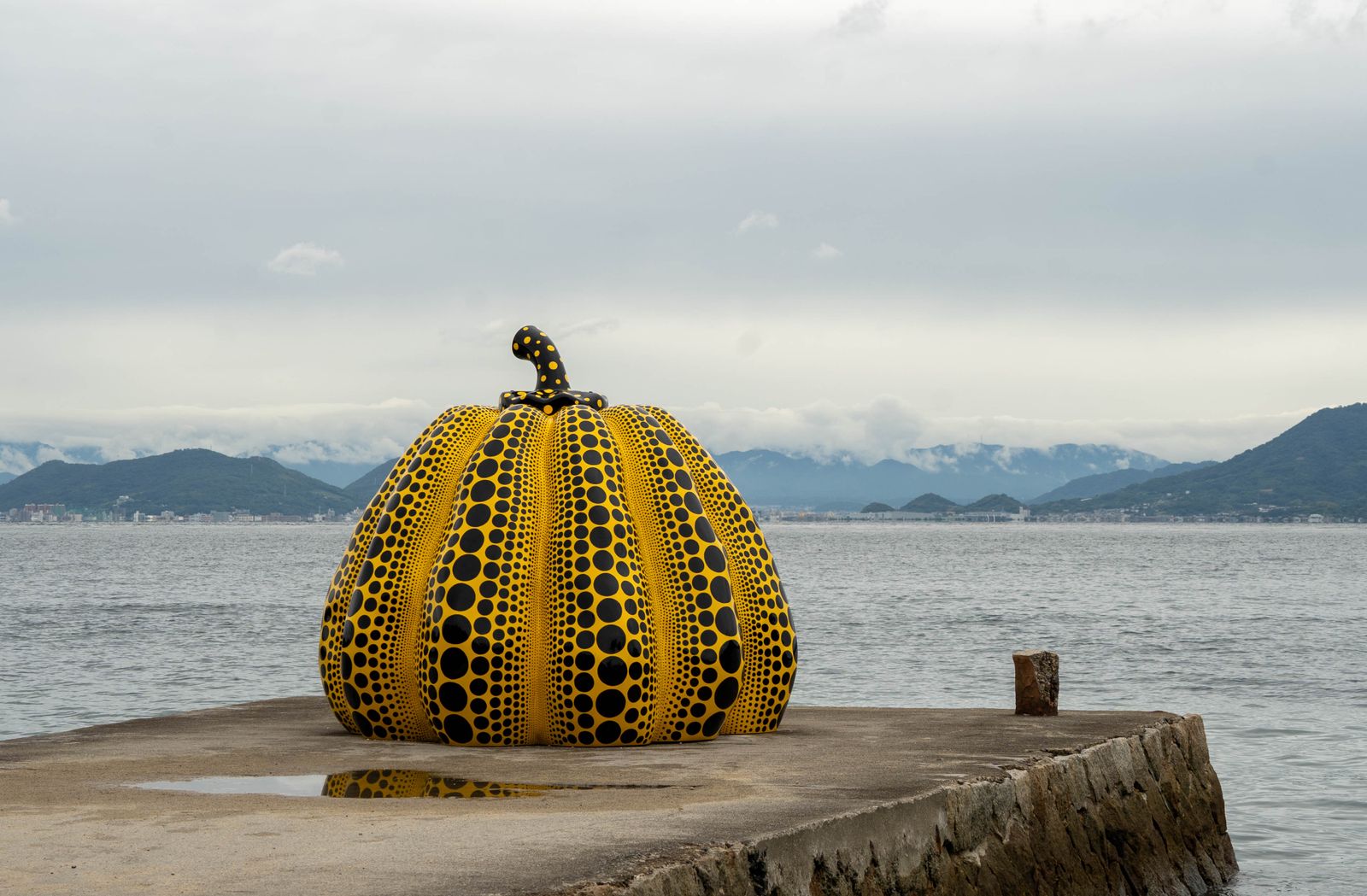 Yayoi Kusama's yellow pumpkin sculpture with black dots on a concrete pier on Naoshima, grey sea and low mountains behind