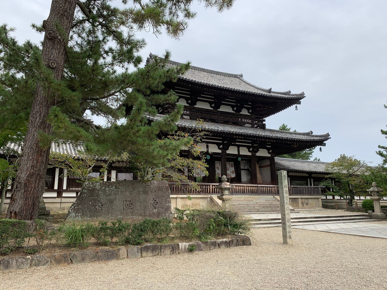 A stone marker inscribed Horyuji in front of a two-storey temple gate, a large pine at the left and a gravel forecourt