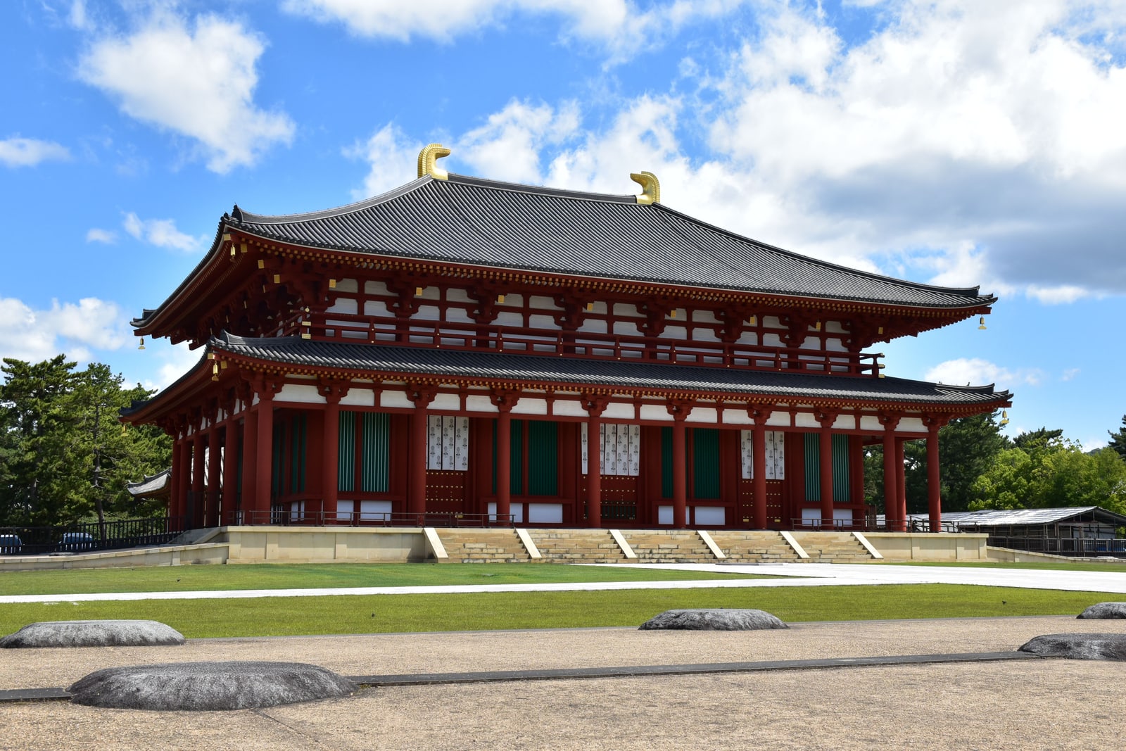 A two-storey red hall with a grey tiled roof and gold ornaments at both ends of the ridge, seen across a lawn under a blue sky