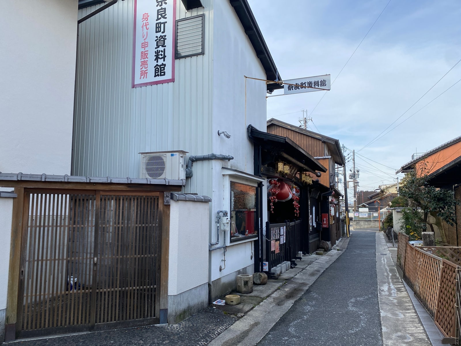 A narrow lane with an old shopfront under a sign reading Naramachi Shiryokan, red cloth charms hung at its door