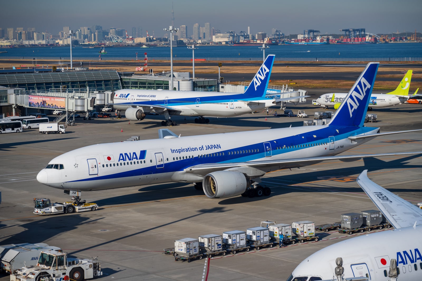 An ANA Boeing 777 on the tarmac at Haneda Airport, with the Tokyo skyline and Tokyo Bay port cranes visible across the water.