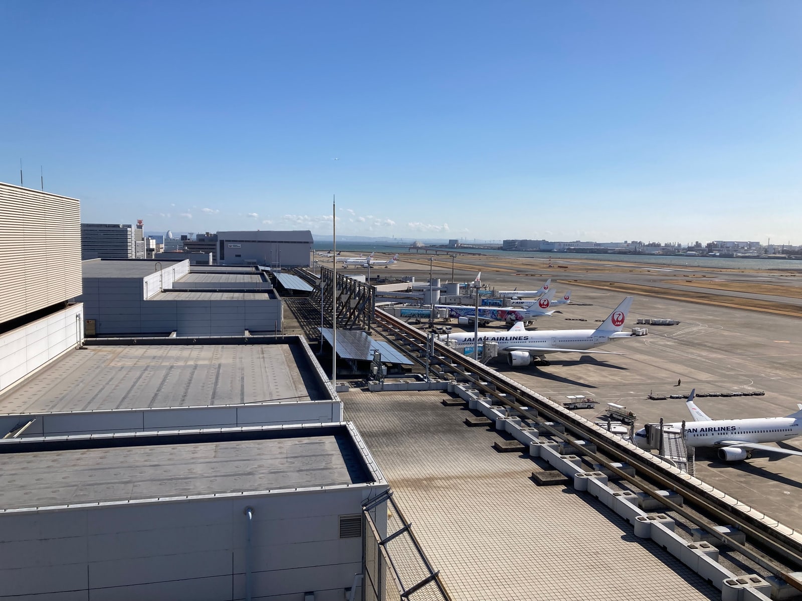 An elevated view over Haneda Airport gates, showing JAL and ANA aircraft parked at jet bridges with Tokyo Bay and the city skyline in the background.