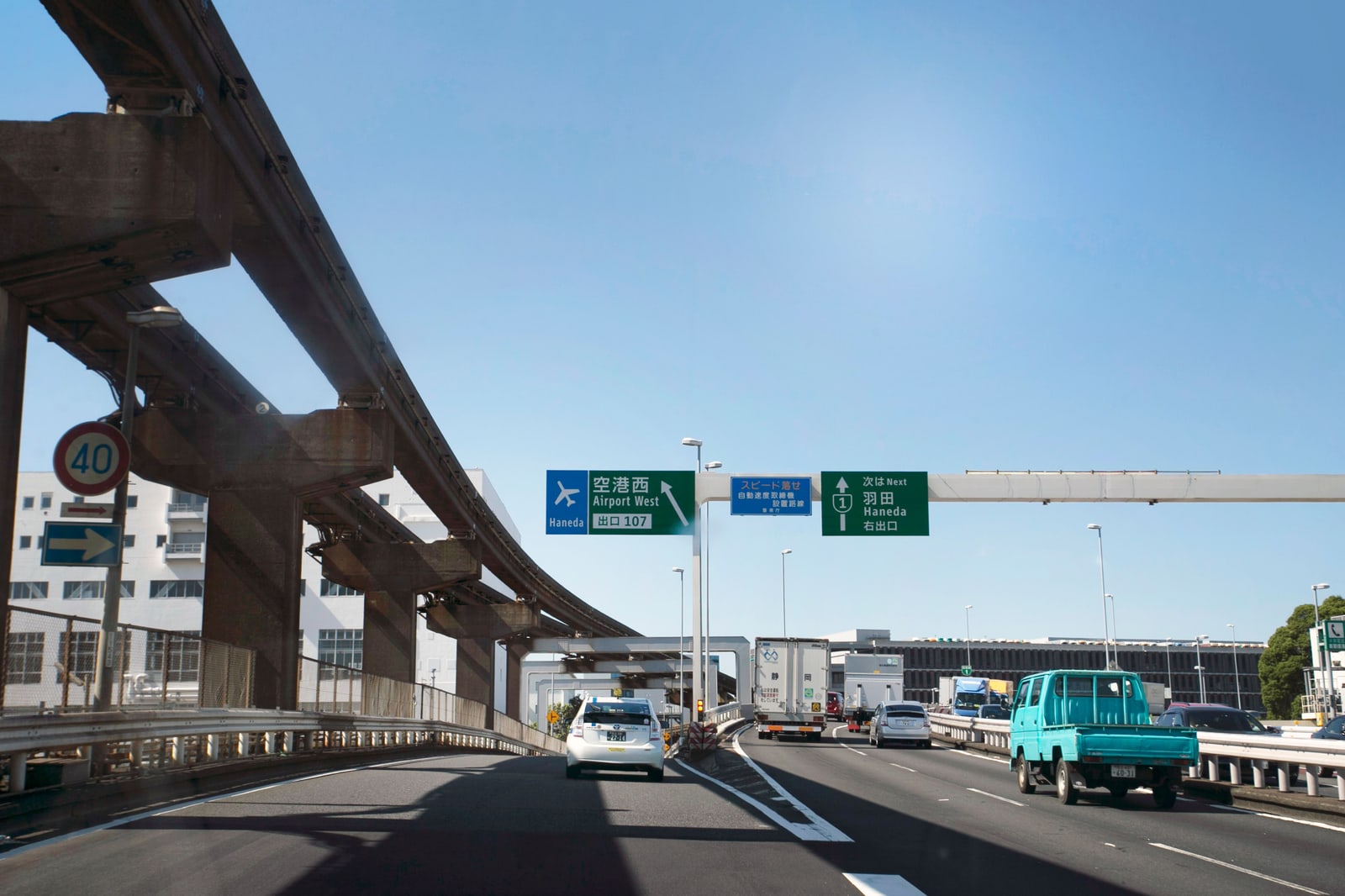 An expressway approaching Haneda Airport, with overhead green directional signs reading Haneda and Airport West, daytime traffic including a taxi and a small truck.