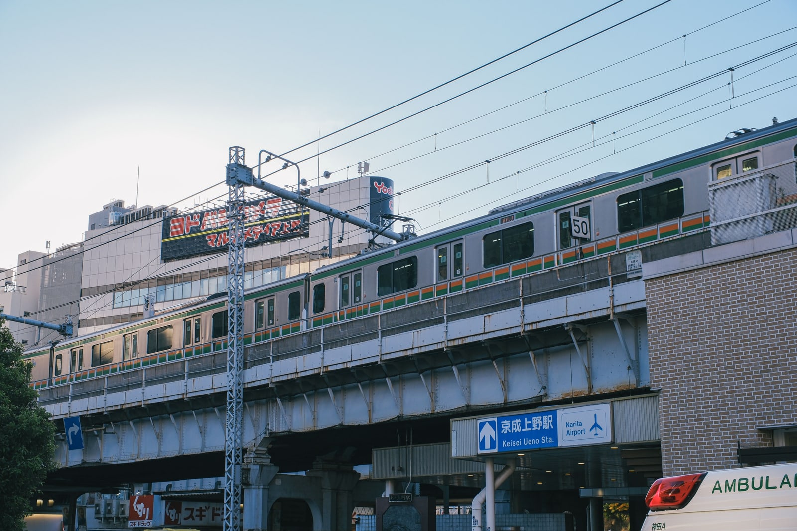 A Keisei train on elevated tracks above a blue sign for Keisei Ueno Station, with an airplane icon and Narita Airport written beneath.