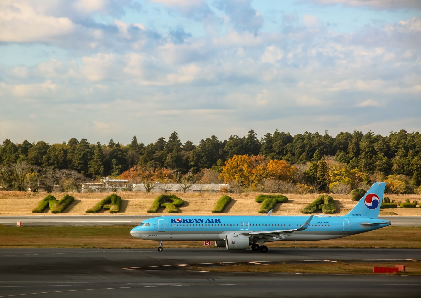 A Korean Air aircraft taxiing at Narita Airport, with hedges spelling NARITA and autumn trees behind the runway.