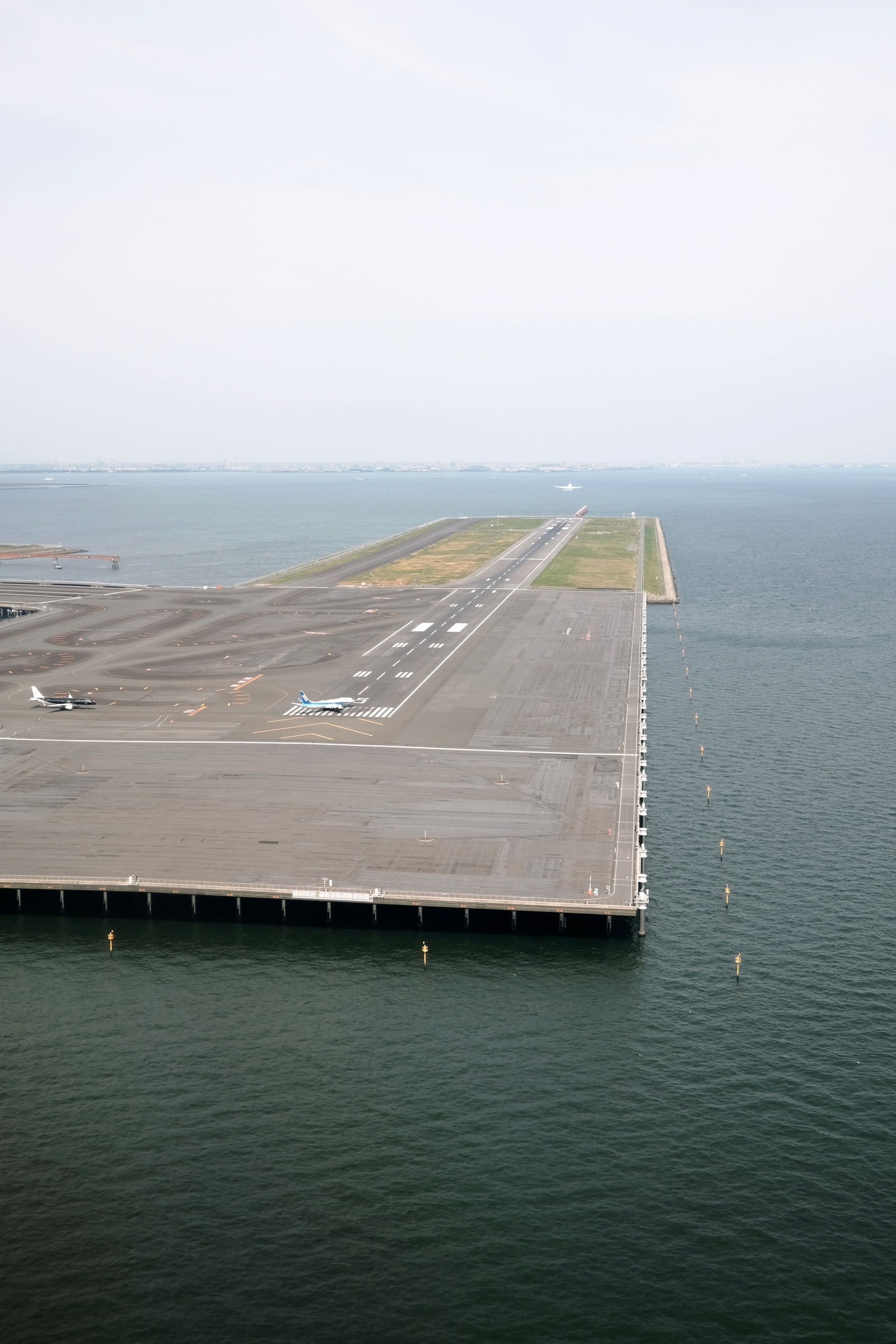 An aerial view of an airport runway built on a pier extending into Tokyo Bay, with two aircraft visible on the tarmac.