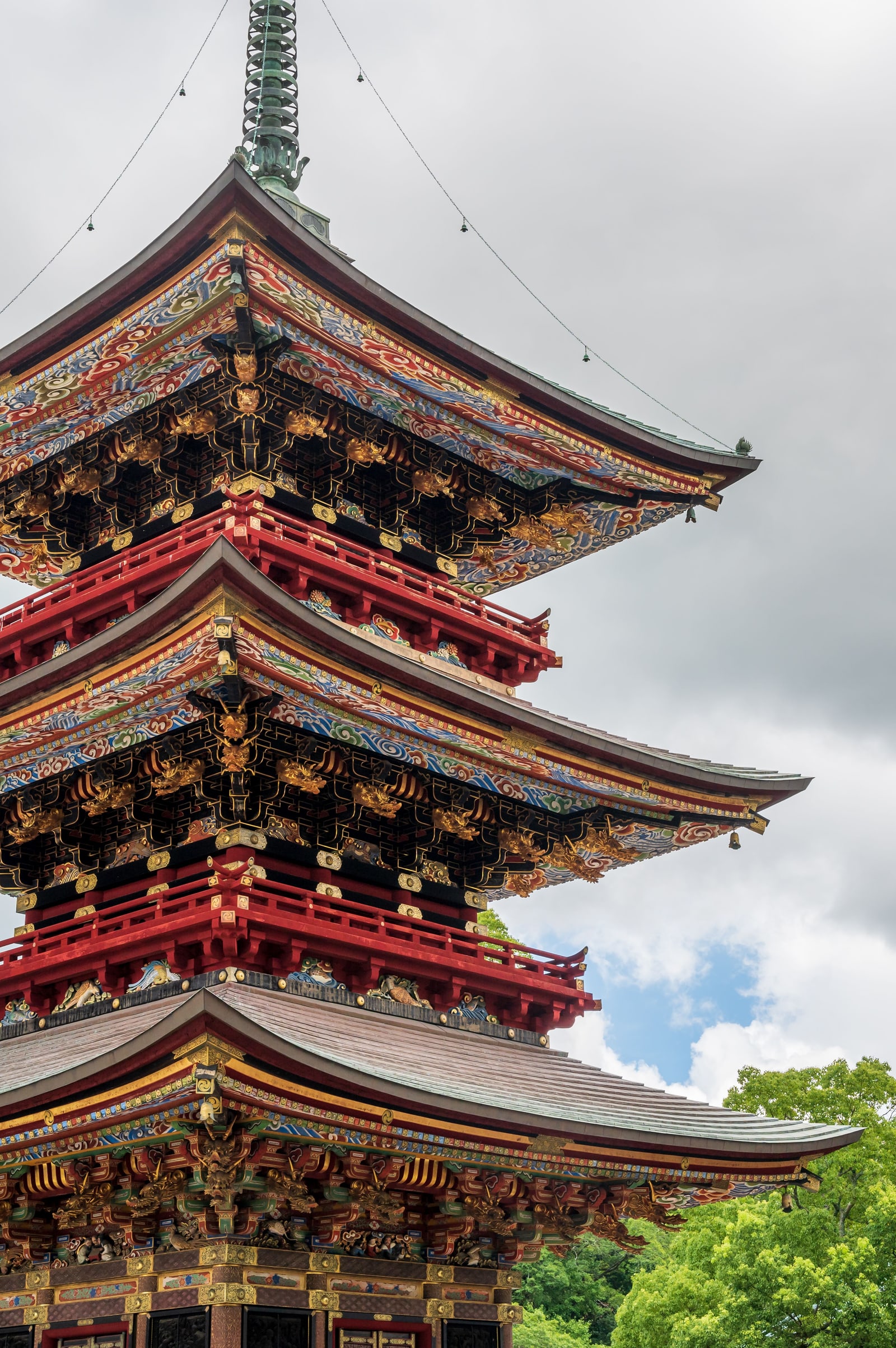 A three-tier red pagoda in Narita with gilded carved brackets and painted eaves under a cloudy sky, green trees at the lower right