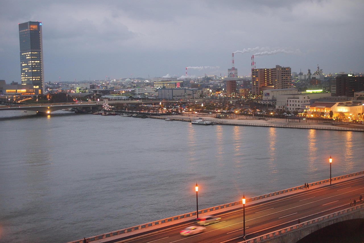 A wide river through the city at dusk, a lit glass tower at left and a lamp-lined bridge in the foreground