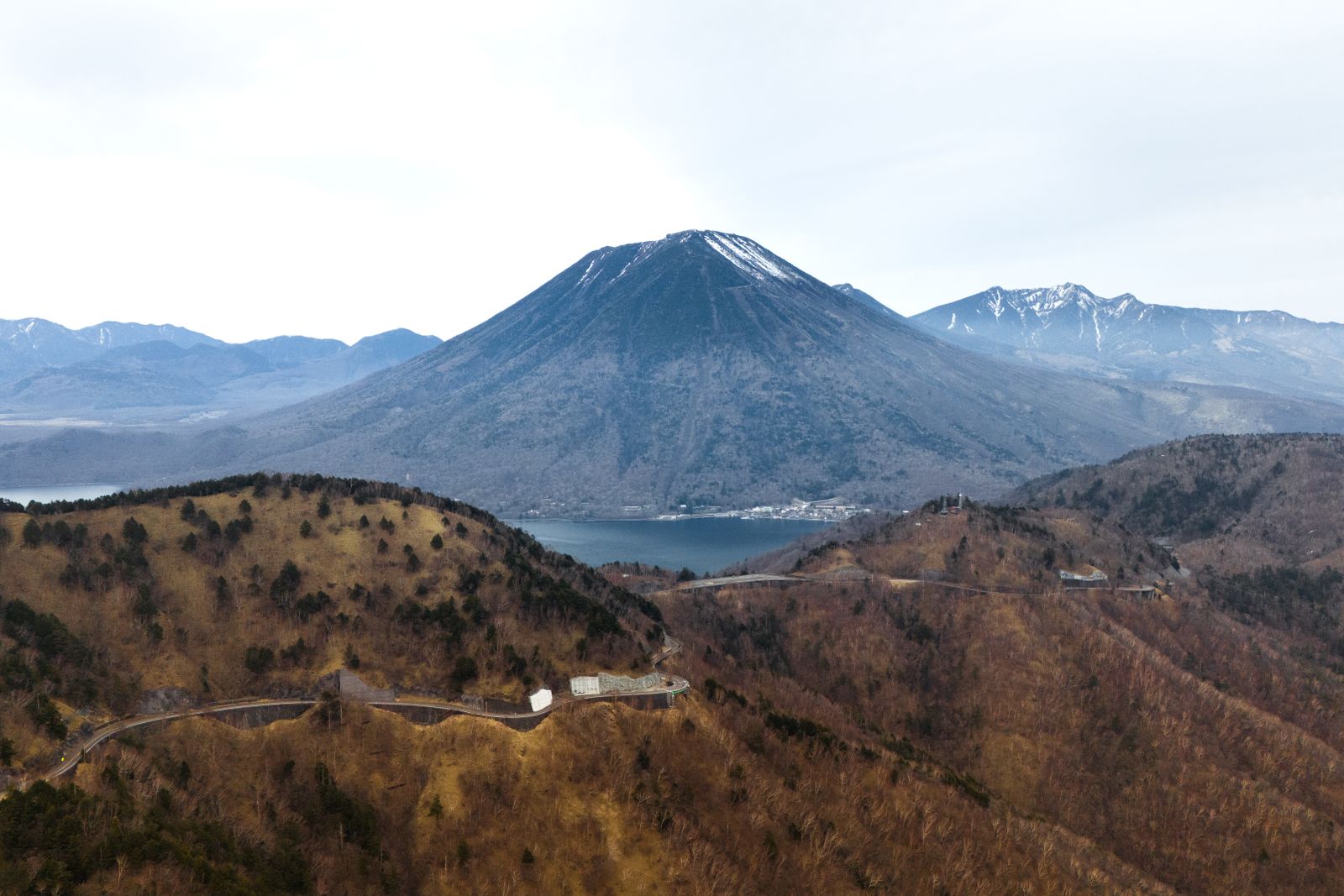 Aerial view of Mount Nantai and Lake Chuzenji, a mountain road winding across the brown autumn slopes in front.