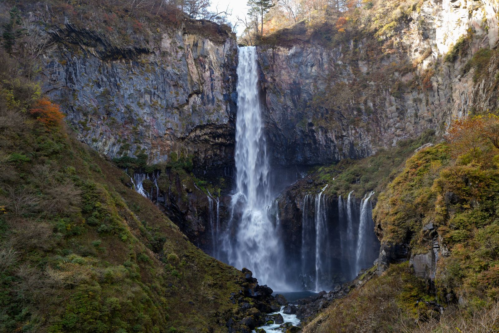 Kegon Falls dropping down a sheer rock wall into a gorge, smaller falls spilling beside it, autumn color on the slopes.