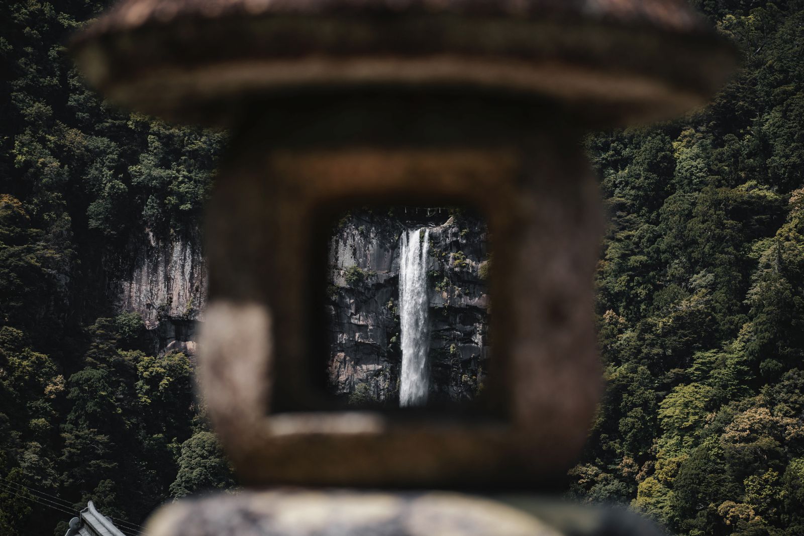 Kegon Falls seen through the window of a stone lantern, dark forest and rock walls around it.