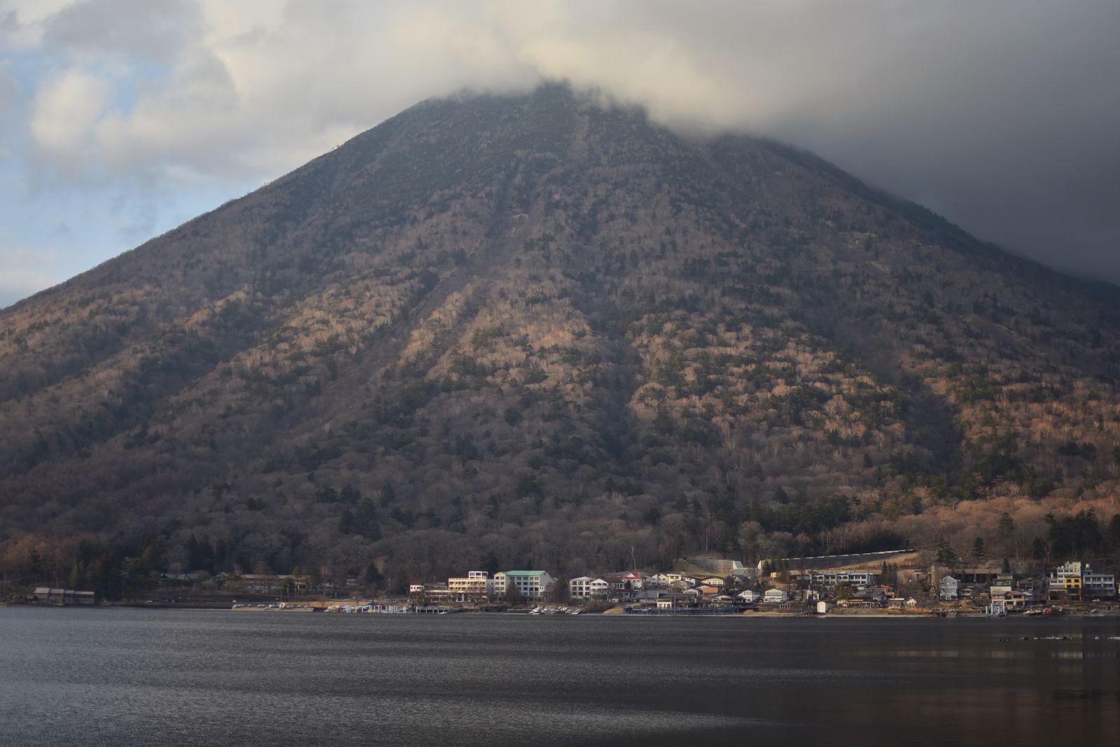 Mount Nantai under low cloud above Lake Chuzenji, a row of lakeside buildings along the far shore.