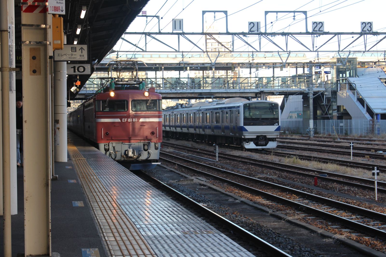A red electric locomotive and a silver JR commuter train at a station, numbered track signs overhead.