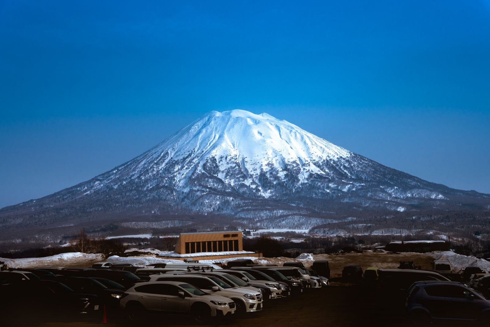 Snow-covered Mt Yotei rising above a car park and a low building in Niseko, Hokkaido, under a deep blue sky