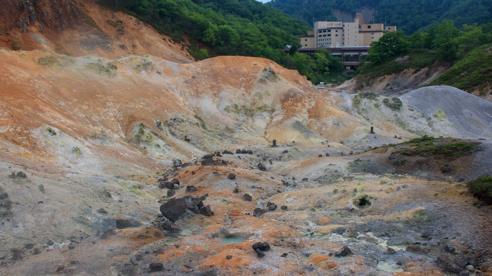 A volcanic valley of orange and grey slopes with a hotel at its head and green hills behind, Noboribetsu Hell Valley