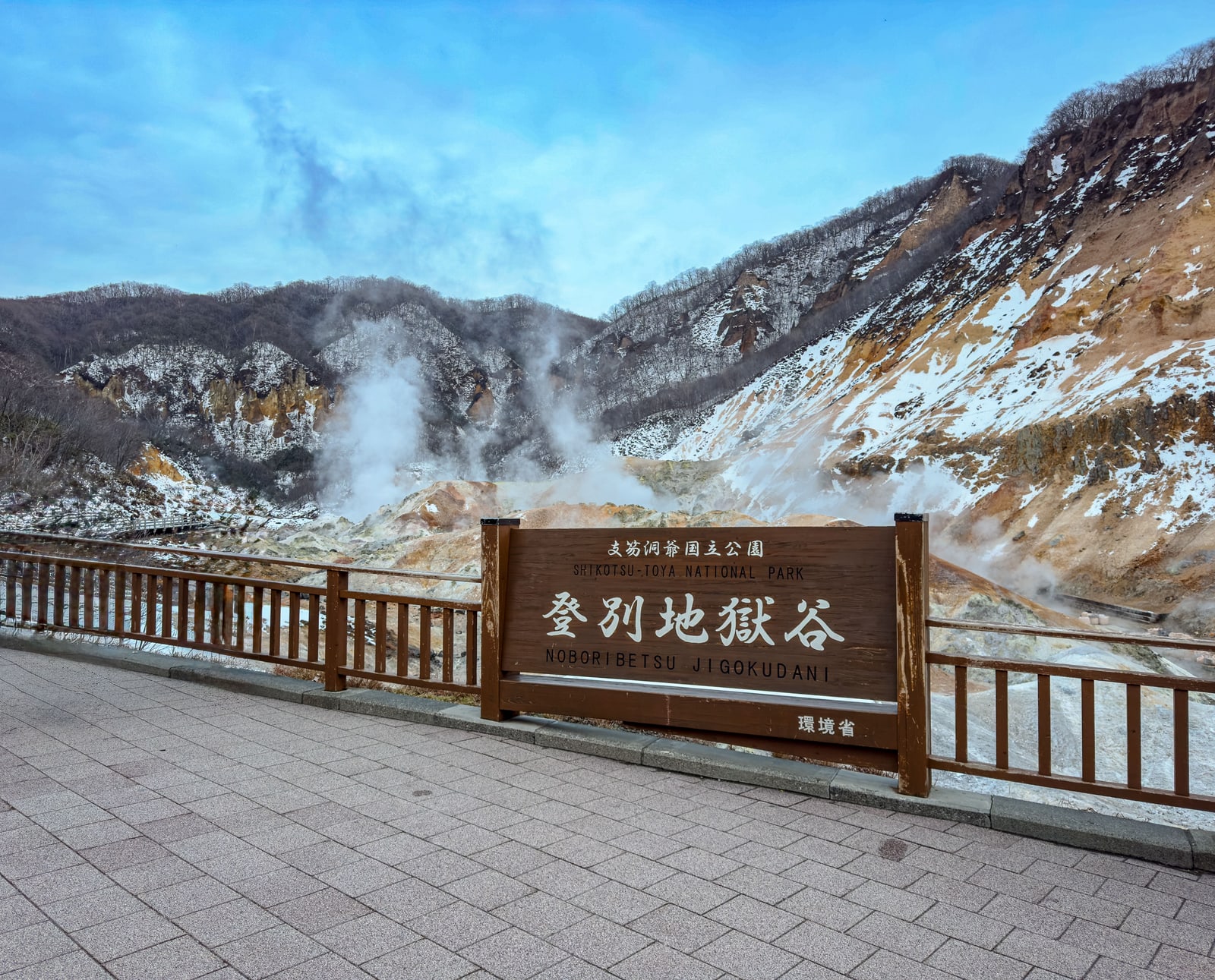 A sign reading Noboribetsu Jigokudani on a railed viewing terrace above a steaming valley of orange rock dusted with snow