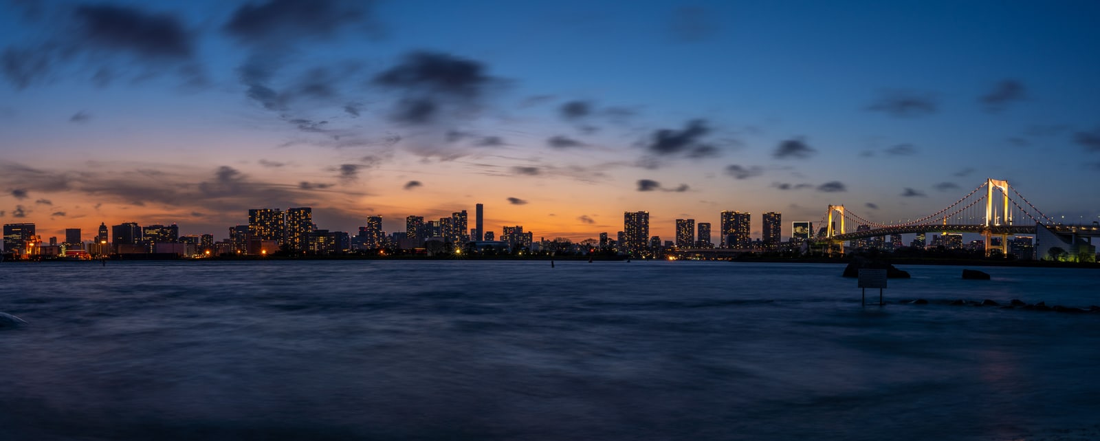 Tokyo Bay at dusk, an orange glow behind the skyline and the lit Rainbow Bridge at the right