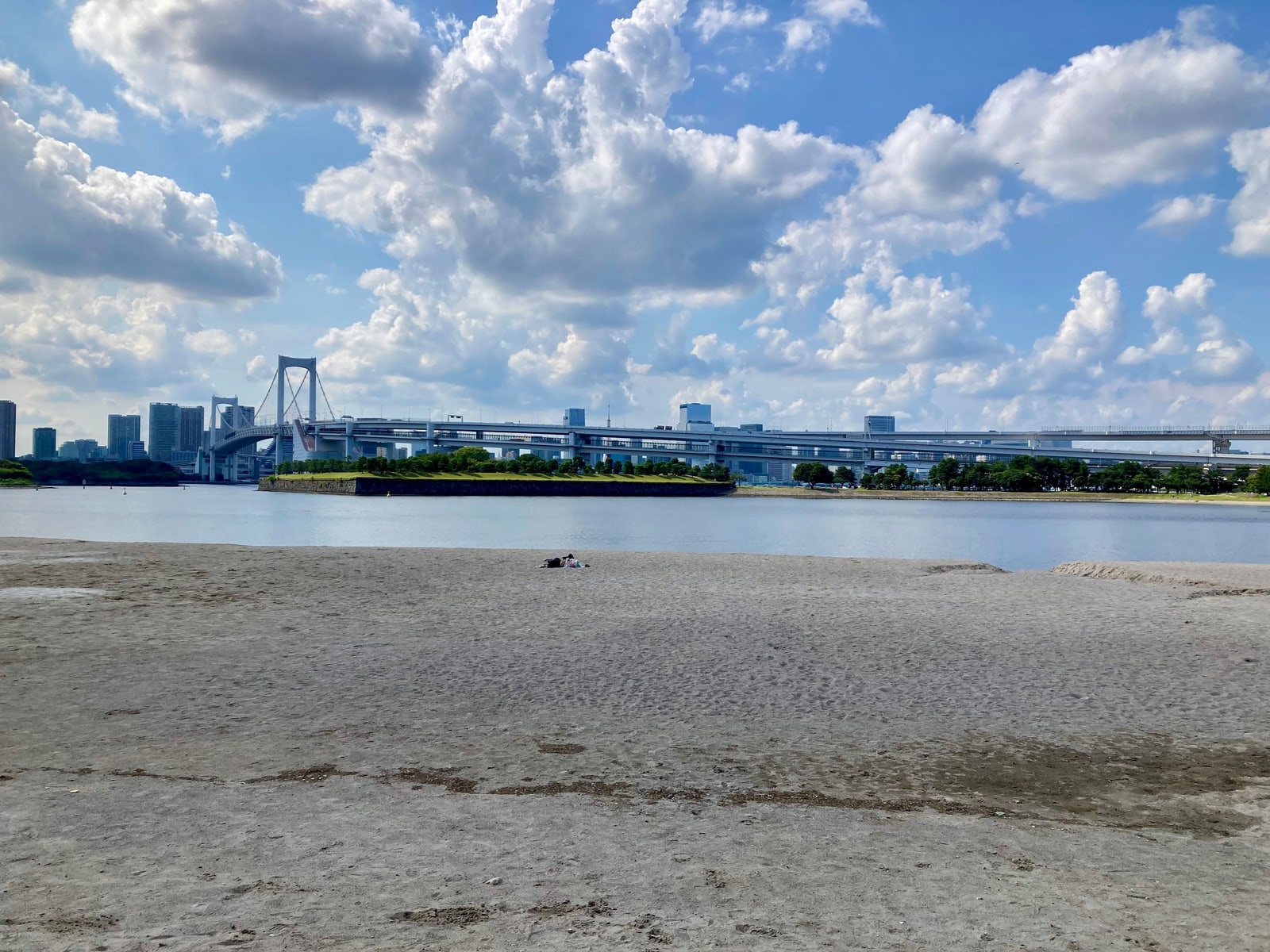 The Rainbow Bridge seen across calm water from the beach at Odaiba, under big white summer clouds