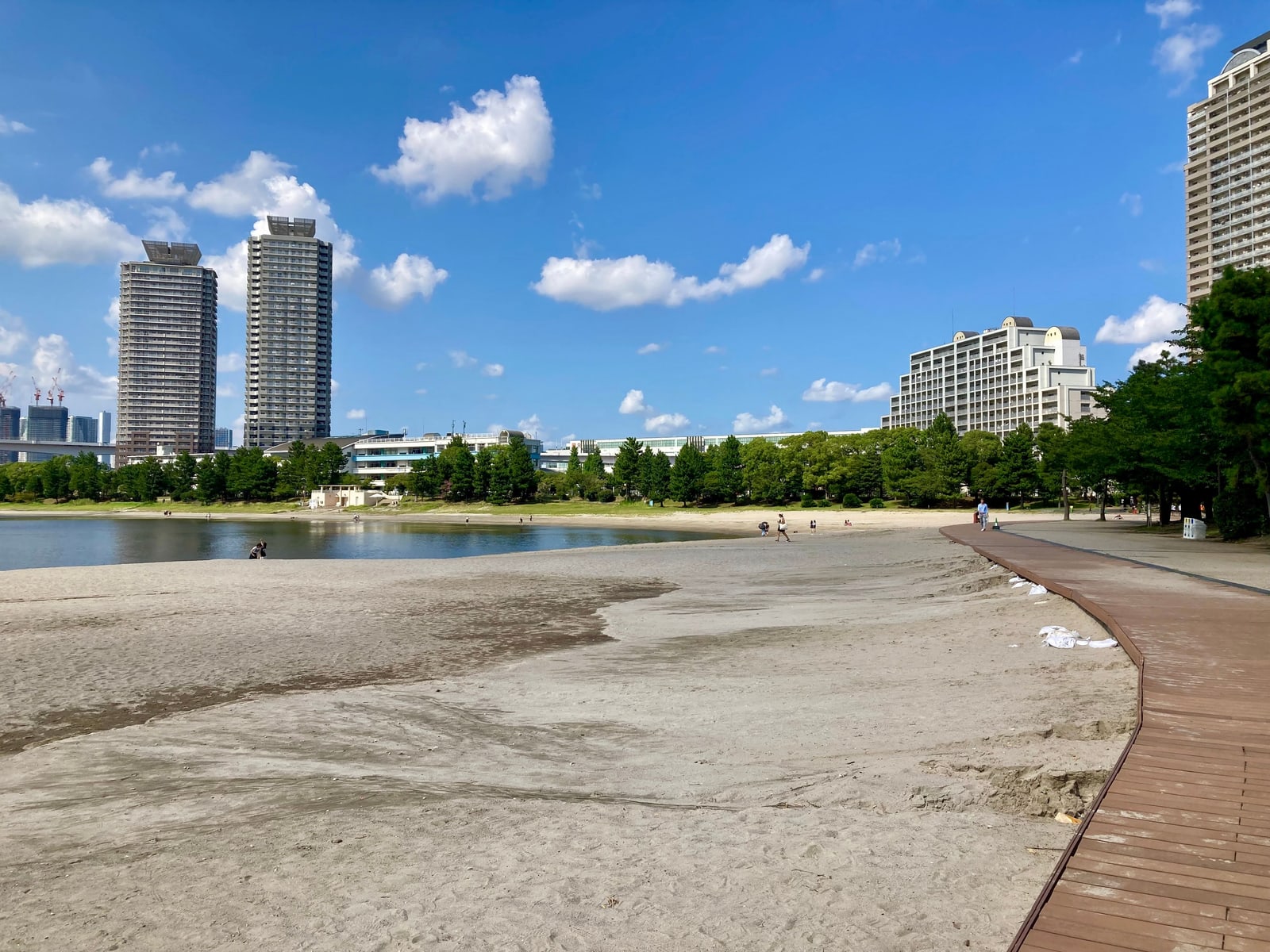 The sand beach and wooden boardwalk at Odaiba under a blue summer sky, residential towers behind the trees
