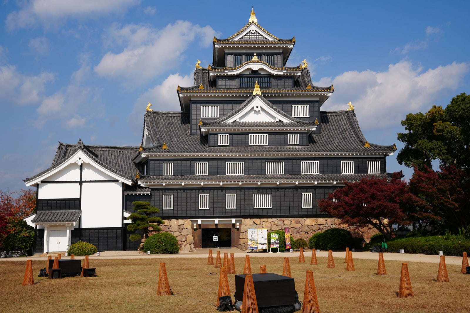 Okayama Castle's black keep with gold roof ornaments, red maples at the side and a lawn set with woven light stands
