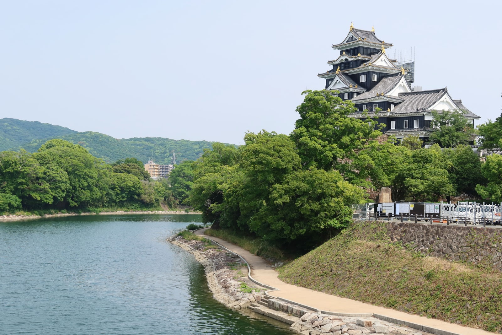 Okayama Castle above summer-green trees, with a river curving away beside a paved bankside path