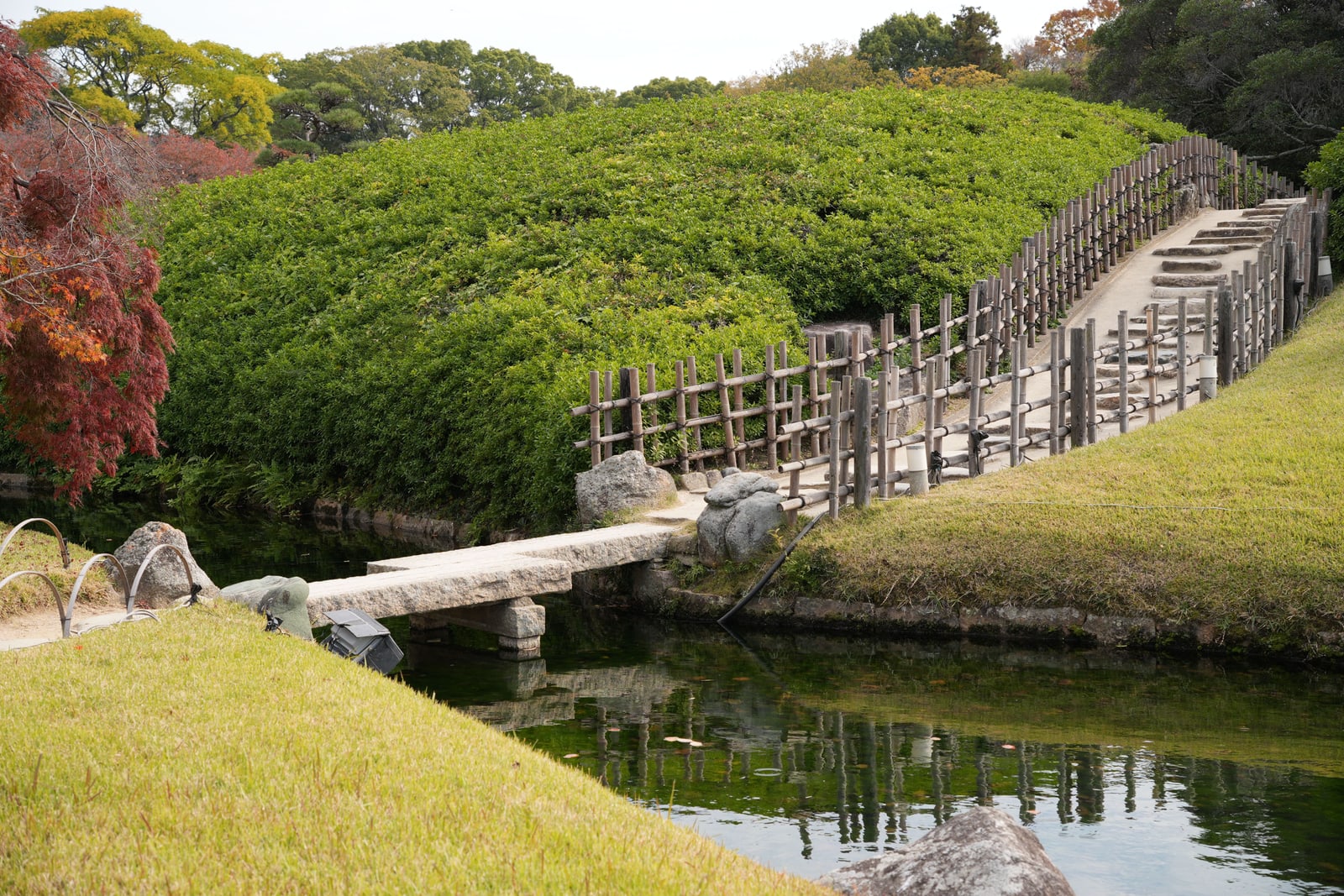 A stone slab bridge over a narrow stream in Korakuen, leading to a stepped path with a bamboo railing beside a clipped green mound