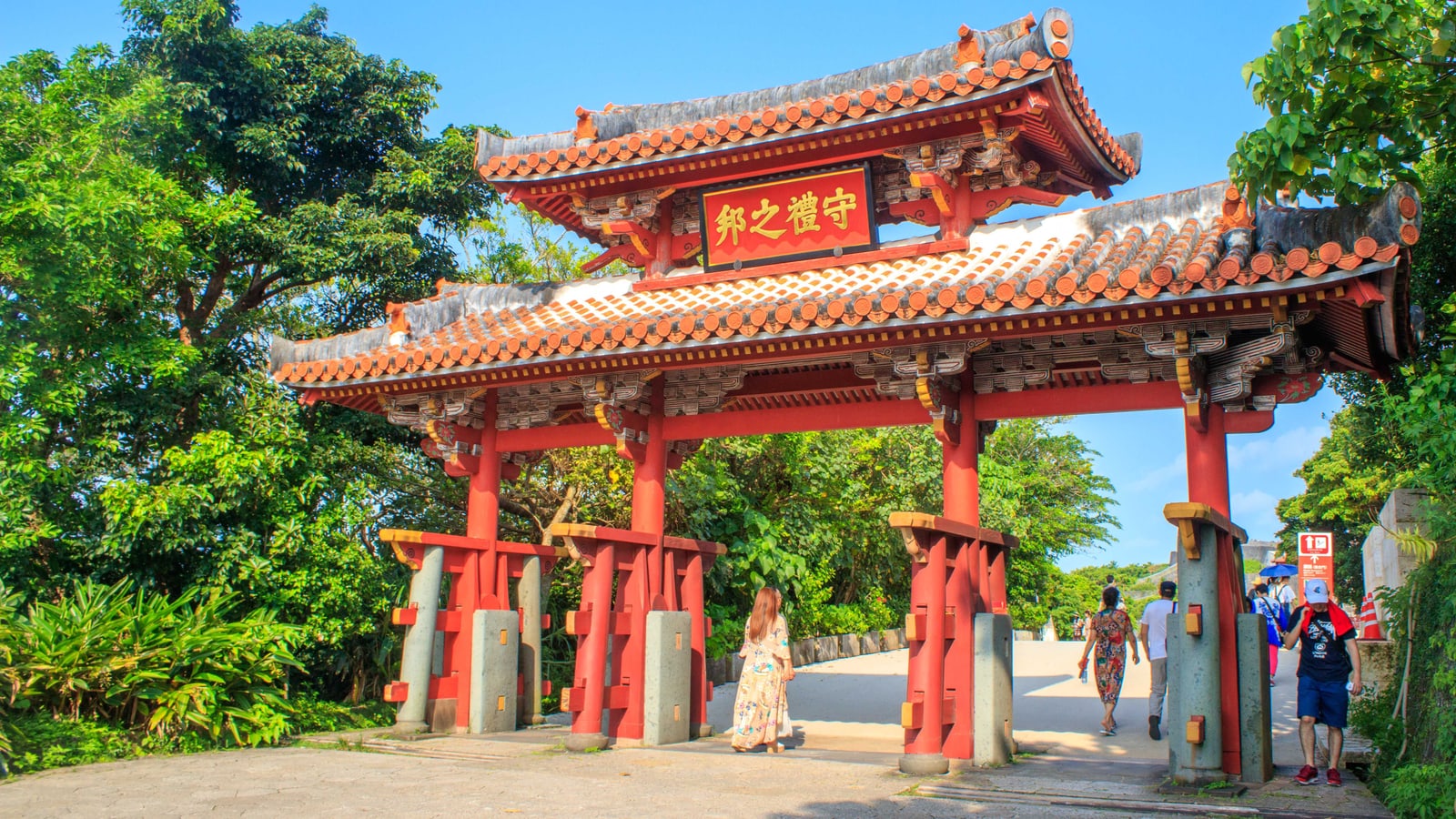 The red Shureimon gate at Shuri Castle Park under a blue sky, trees on both sides