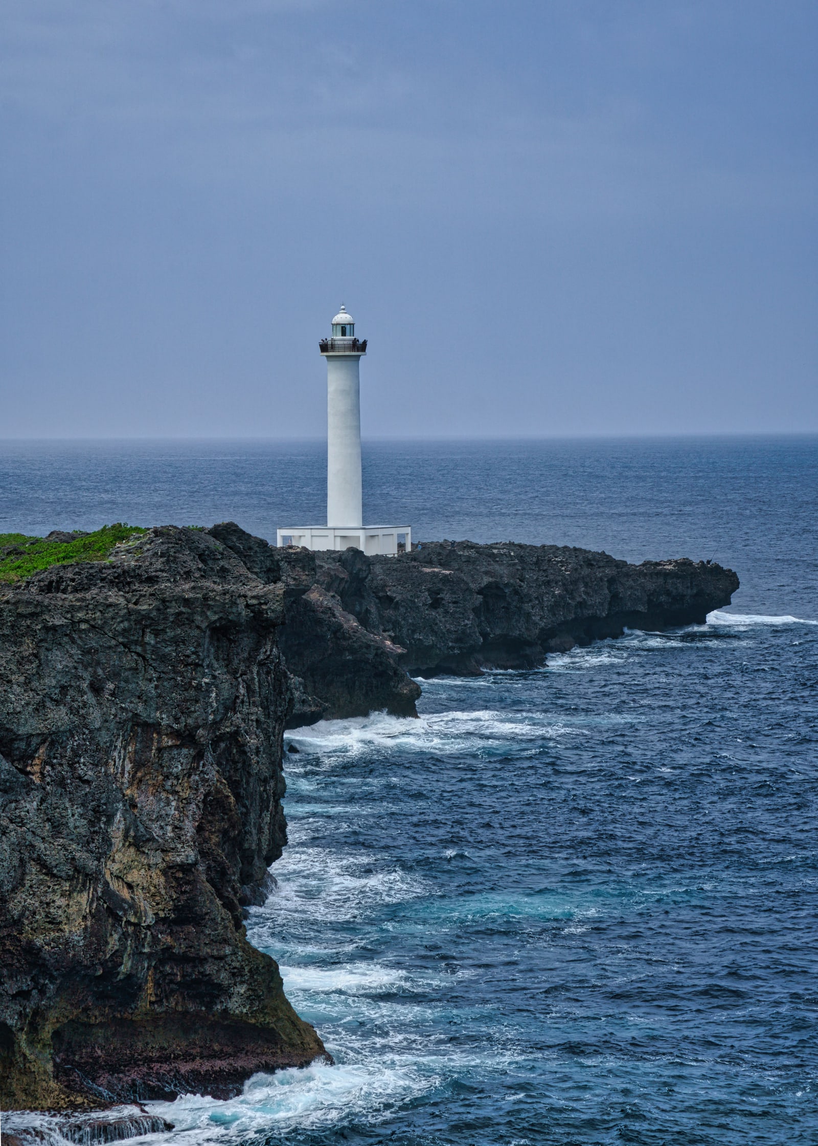 The white lighthouse at Cape Zanpa on a dark limestone headland, with surf below under an overcast sky