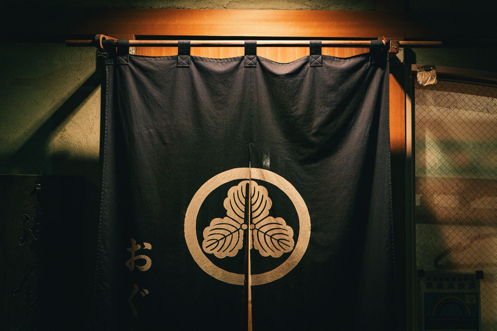 A dark split noren curtain carrying a family crest, hanging in a lit doorway with Japanese characters beside it.