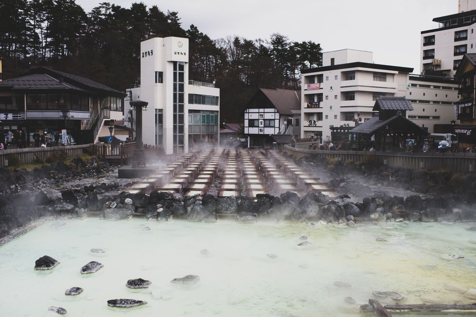 A hot-spring town's yubatake, wooden channels cooling the source water with steam drifting across the basin.