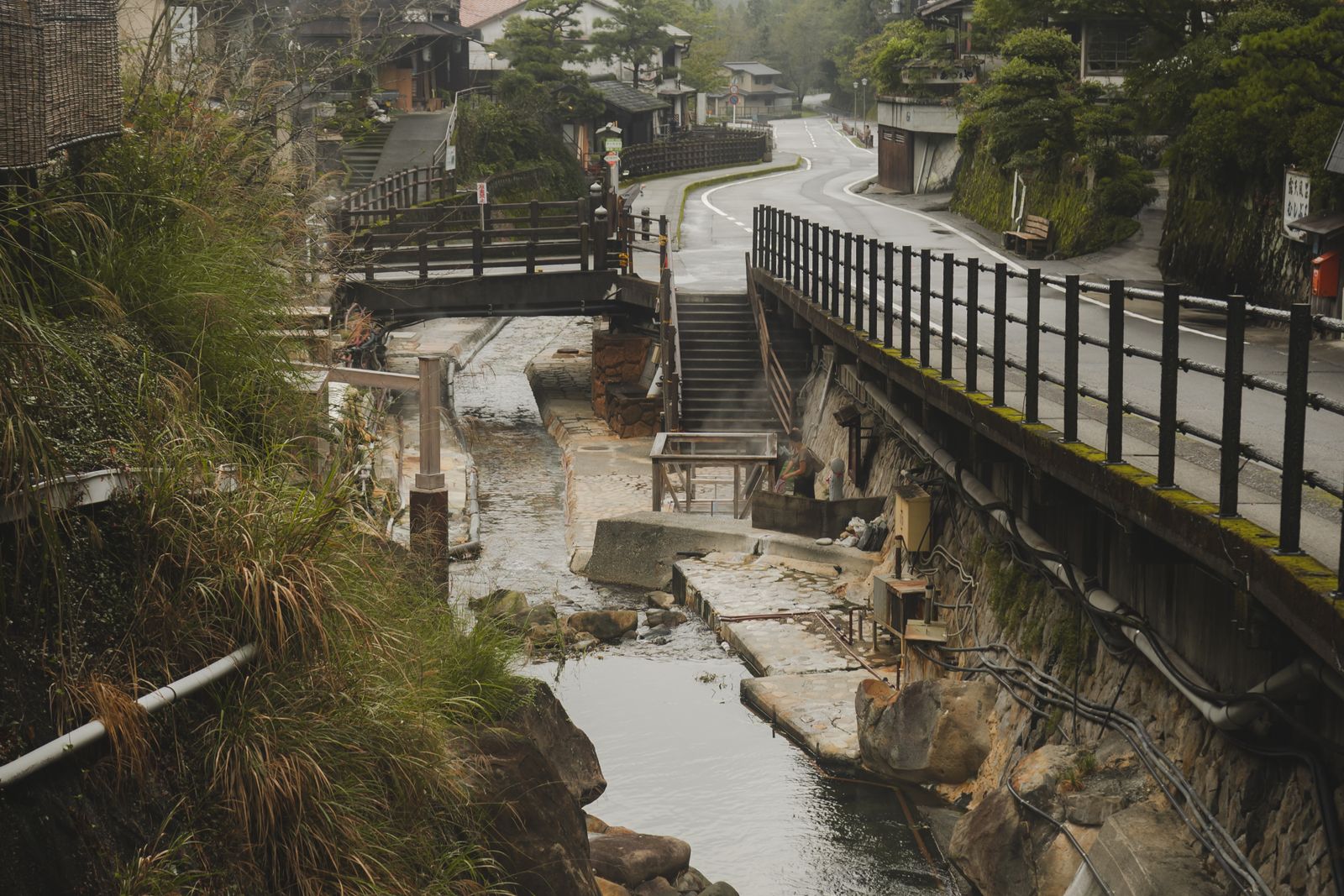 A stream running through an onsen village, a bridge and stone stairs, steam rising at a hot-spring basin, one figure in the distance.