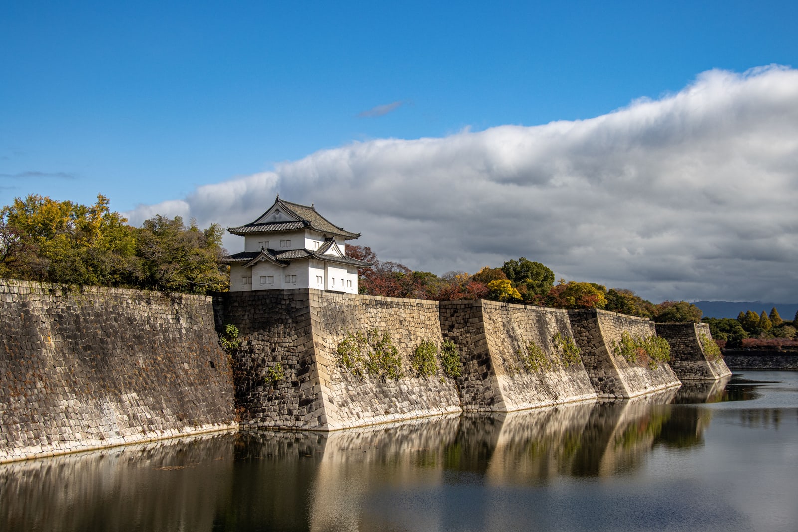 Osaka Castle's outer stone walls and a corner turret reflected in the moat under a bright sky.