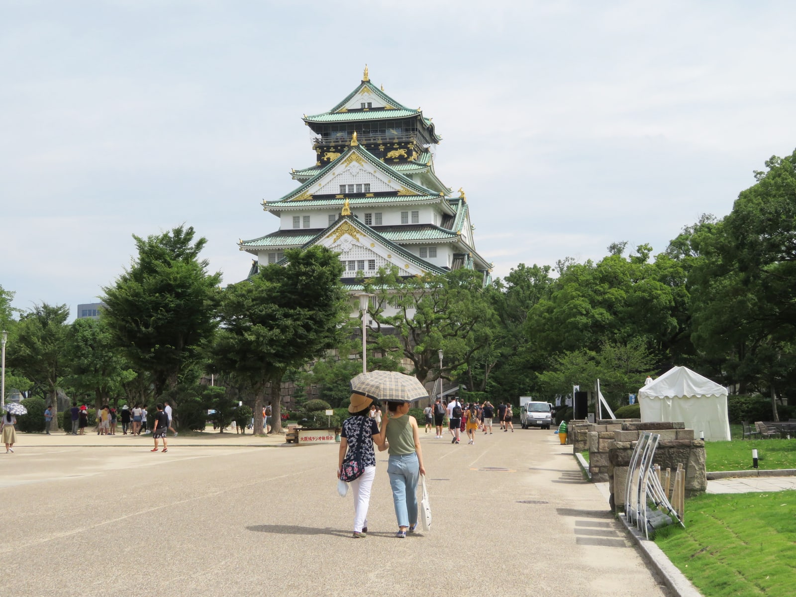 Osaka Castle's main tower at the end of a wide paved approach, two visitors under a parasol walking toward it.