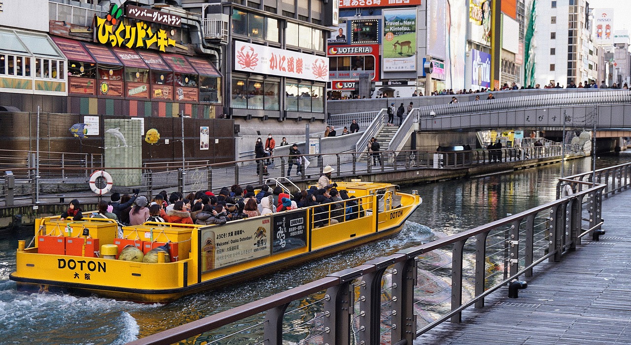 An open yellow sightseeing boat full of passengers on the Dotonbori canal, with the riverside walk and shop signs behind.