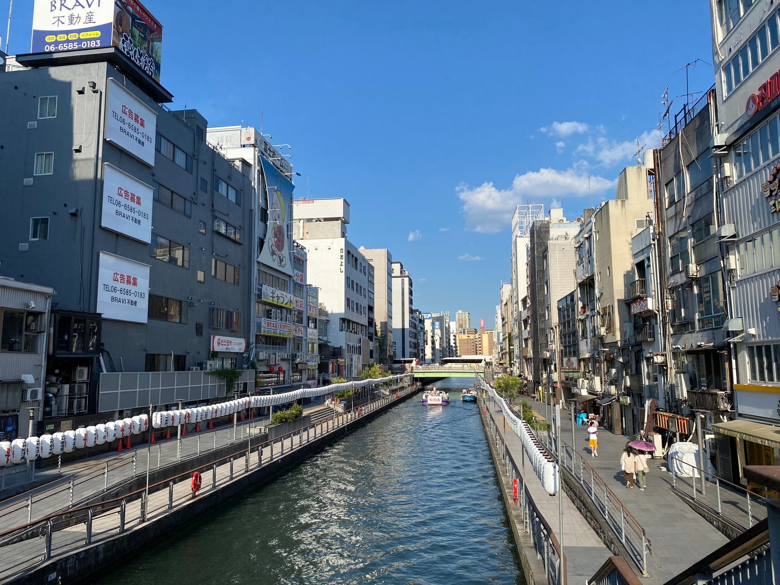 The Dotonbori canal in daylight, with unlit signs, a paved riverside walk and few people.