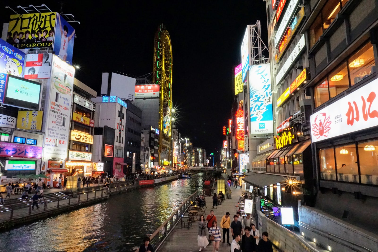 The Dotonbori riverside walk at night with people, lit signs, and a yellow oval Ferris wheel on a building to the left.