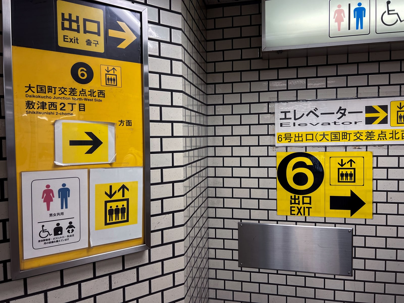 Yellow exit signs on a white tiled wall inside an Osaka Metro station, with a numbered exit and an elevator arrow.