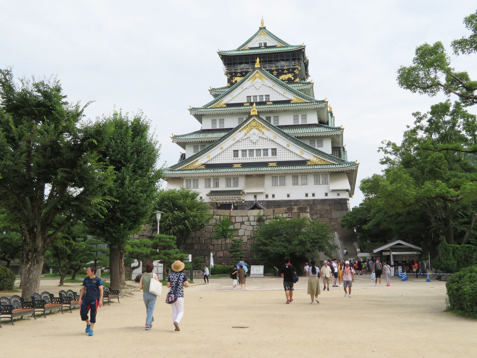 The main tower above a wide sandy square, a scattering of visitors walking toward it between tall trees.