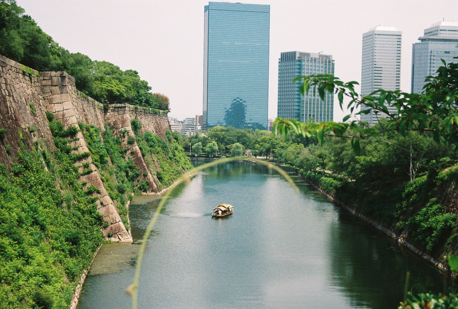 A small roofed boat on a green moat between a steep stone wall and trees, glass office towers behind.
