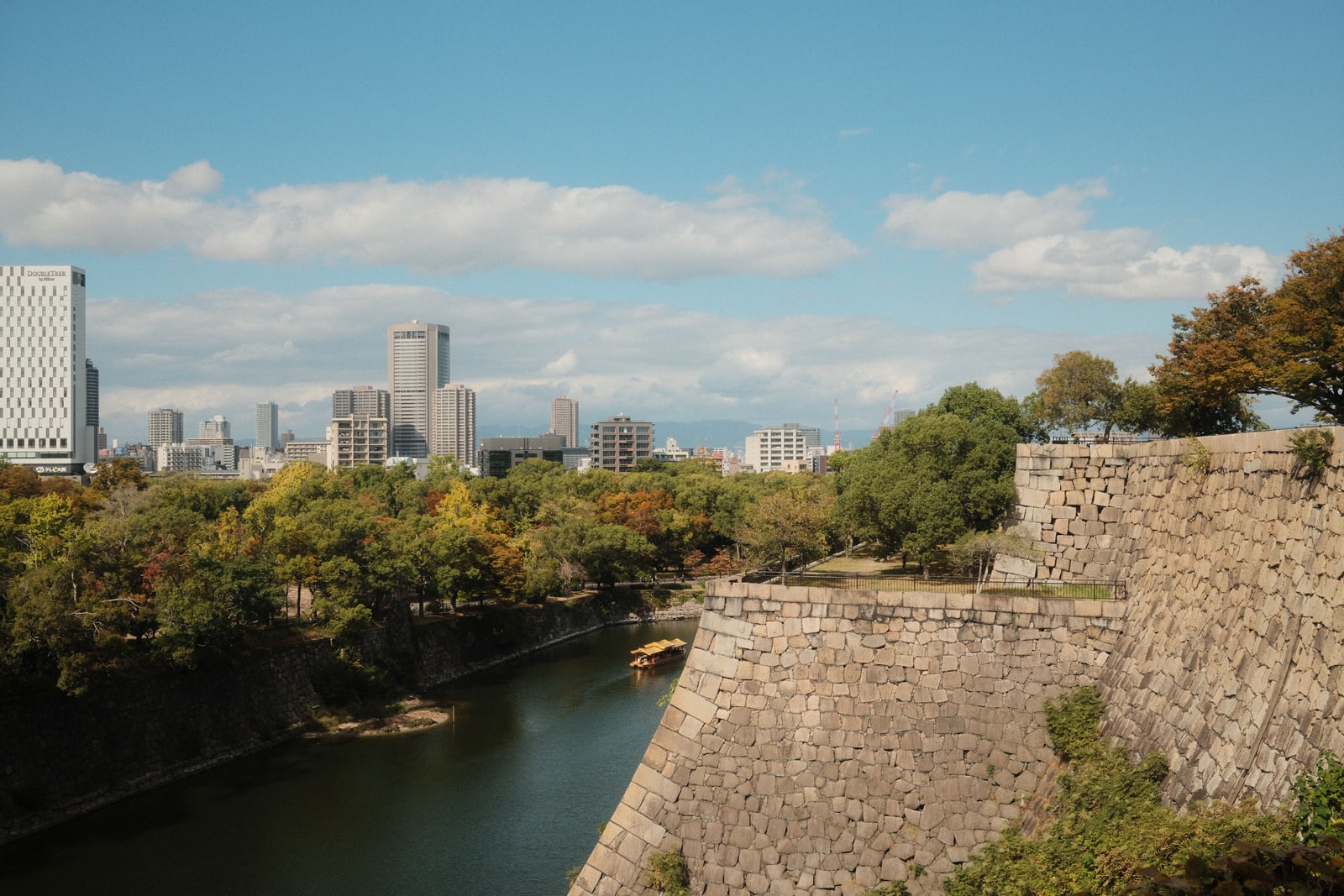 A curving moat below high stone walls and autumn trees, office towers on the skyline.