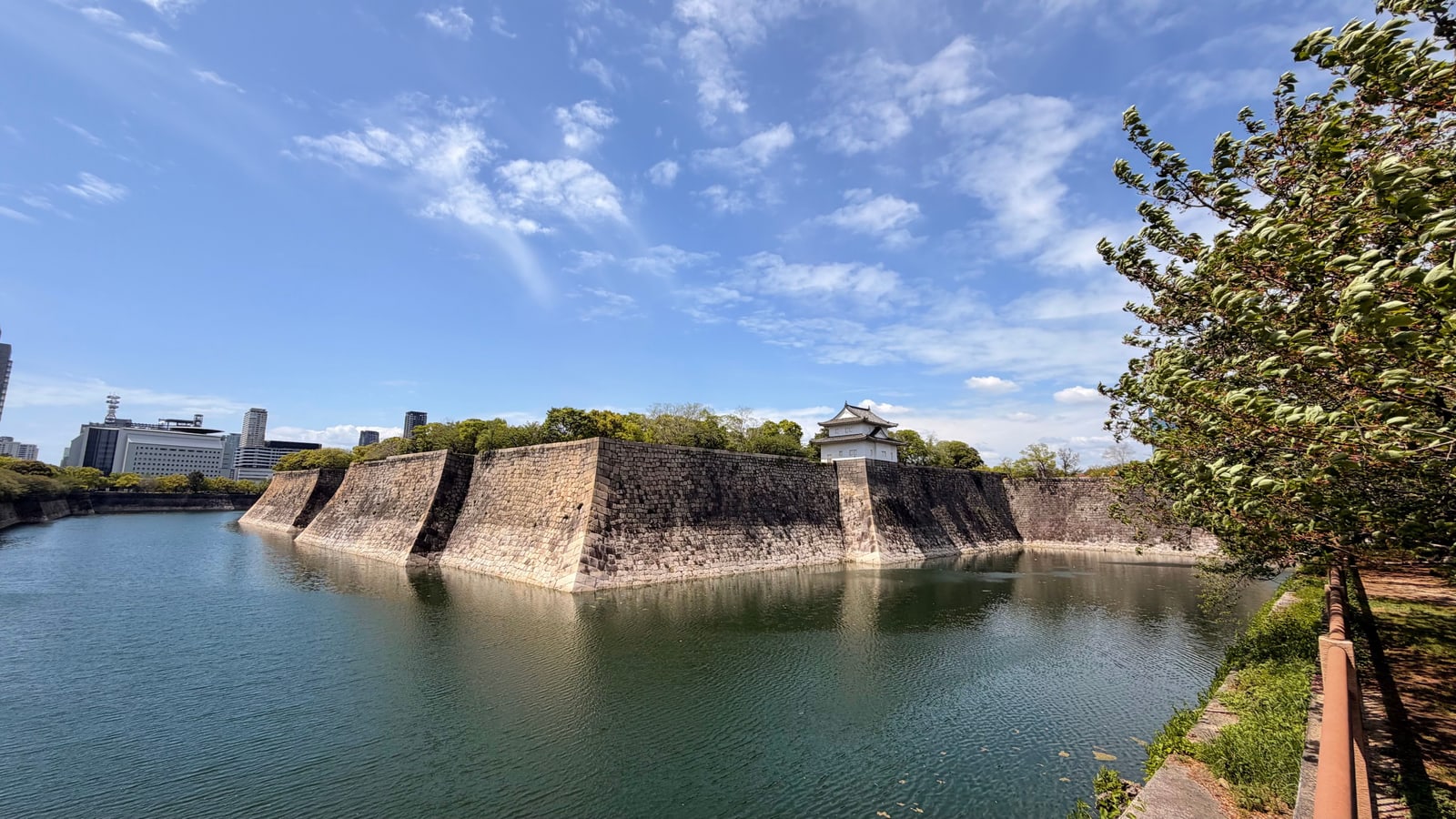 A wide moat below long angled stone walls with a small corner turret, office buildings far off to the left.