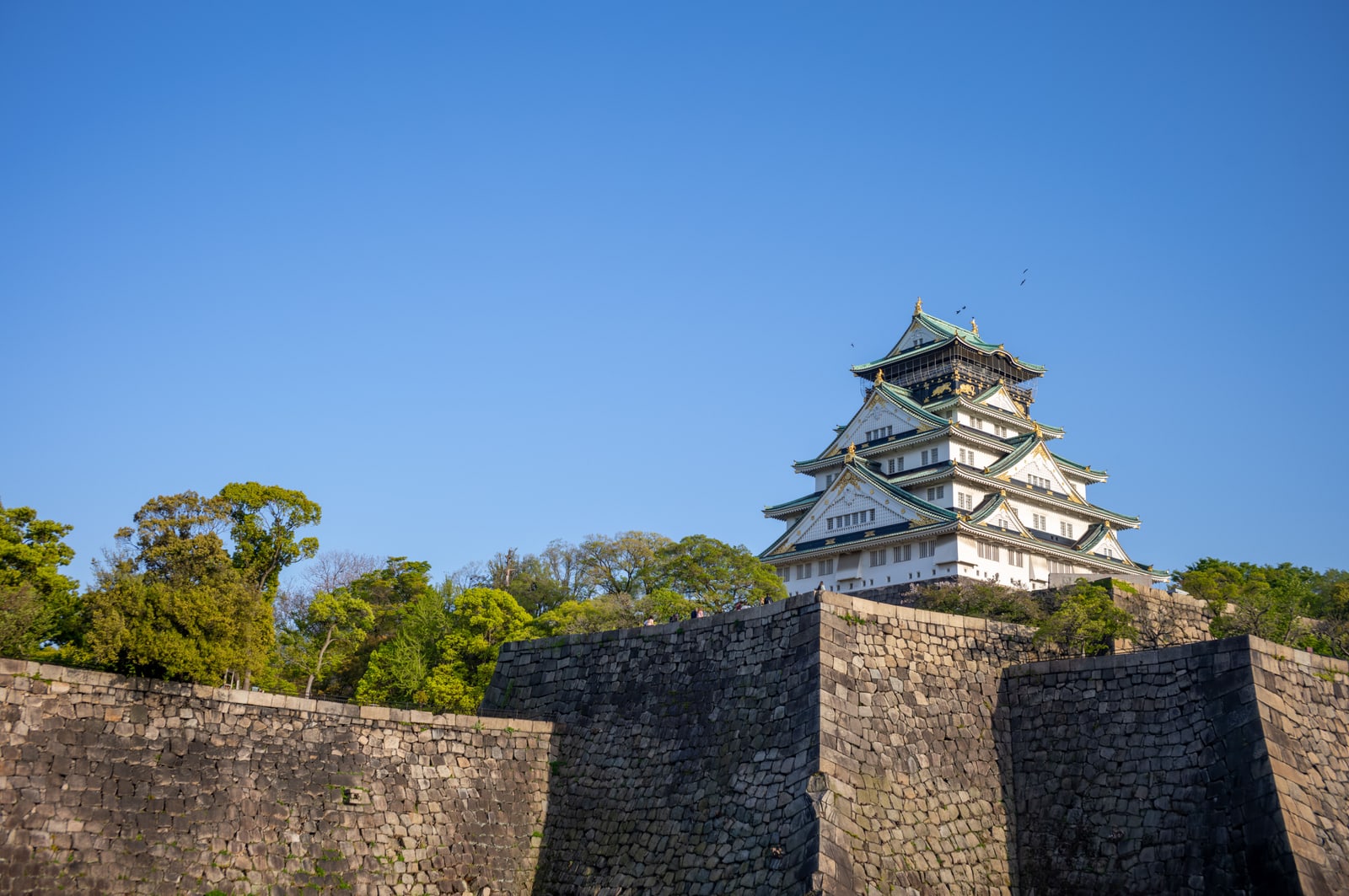 Osaka Castle's main tower rising behind tall dark stone walls under a deep blue sky.